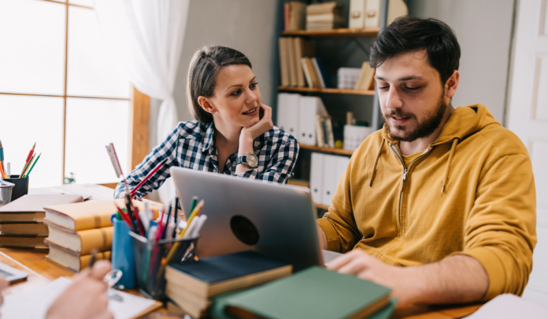 A man and woman talk while he uses a laptop.