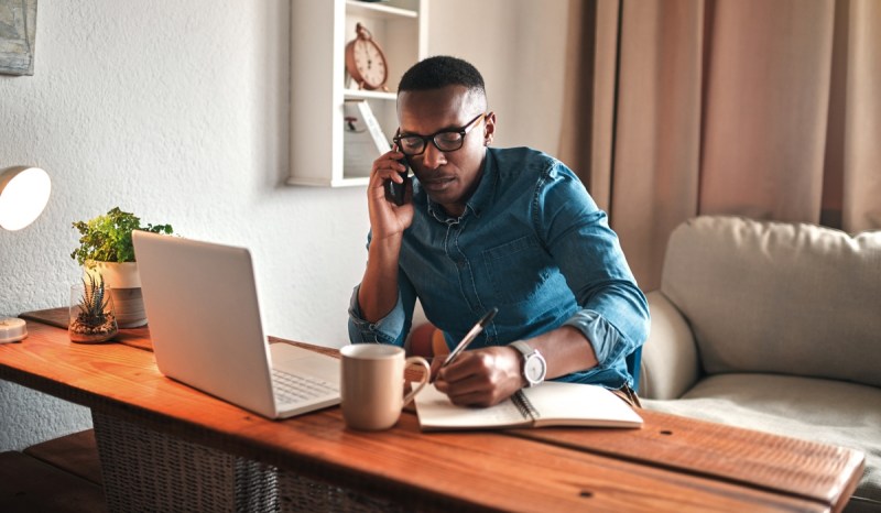 Man sitting at desk with laptop, writing in a notebook with a pen