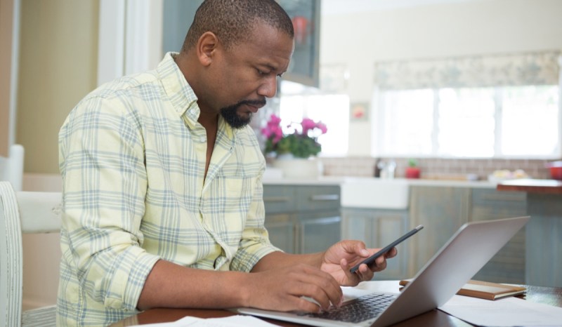 Man working on laptop while looking at a smartphone.