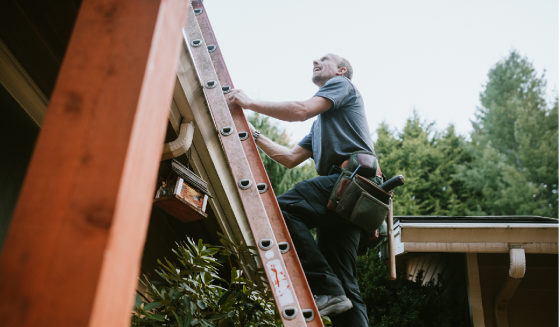 A man in a t-shirt climbs a ladder on the side of a house.