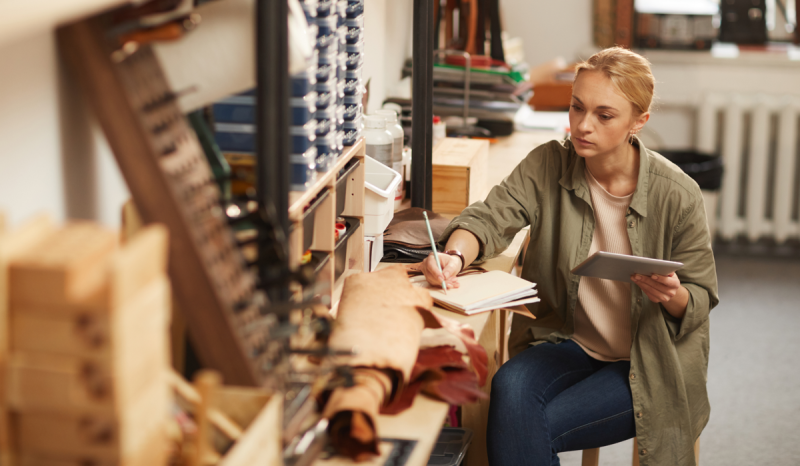 Woman at workbench holding one notebook while writing in another.