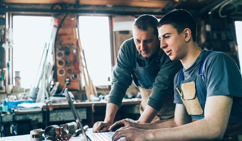 Two men, one sitting and one standing, looking at a laptop that's on top of a work bench, in a studio.