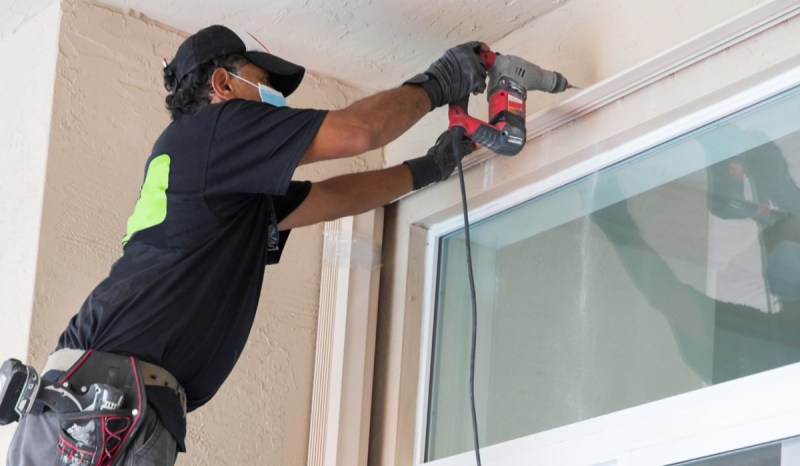 Man wearing baseball cap drilling above a window.