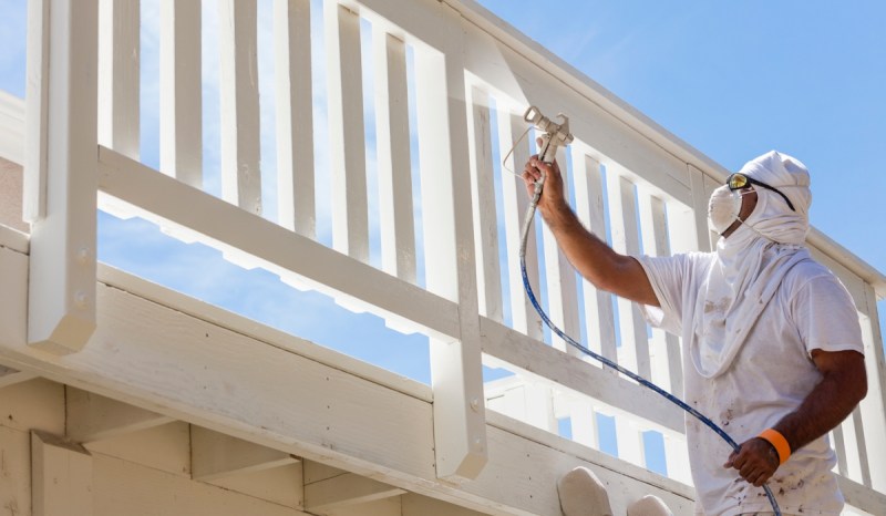 Person wearing all white and a mask while spray painting a fence white.