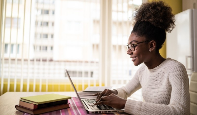 Woman with glasses typing on laptop. Three books in front of laptop.