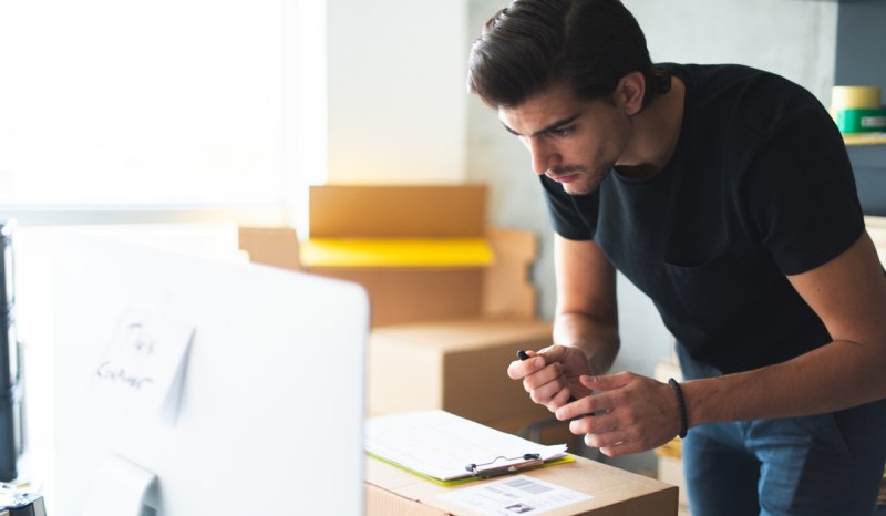 Man holding pen while bending over desk and looking at computer monitor.