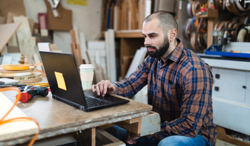 Man wearing plaid shirt, sitting in workshop while working on a black laptop.