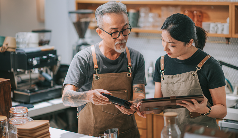 Man and woman working in a coffee shop showing each other something on their devices.