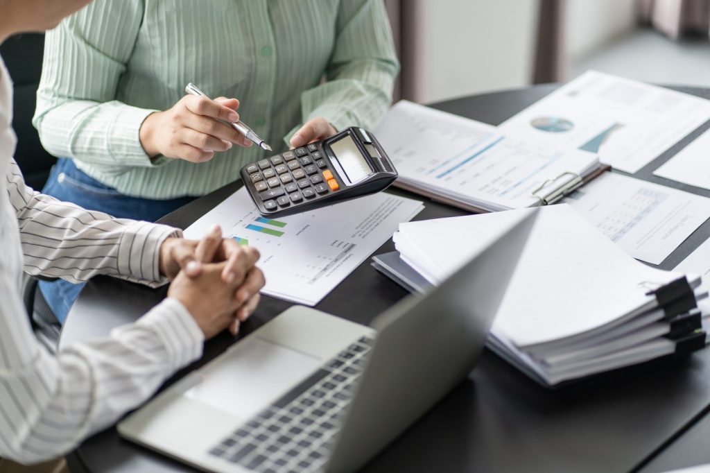 Two people sitting at a table covered with documents while using a laptop and calculator